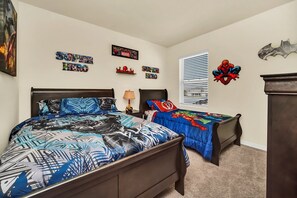 Une chambre avec deux lits, une fenêtre avec des stores et des éléments de décoration murale sur le thème des super-héros.