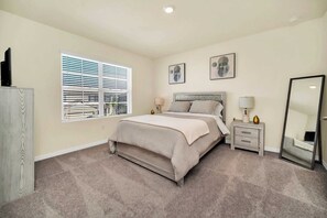 Un dormitorio con cama, dos mesitas de noche, cómoda, ventana con persianas y dos cuadros colgados en la pared.