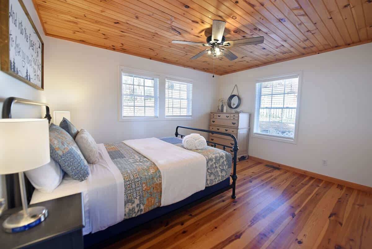 A bedroom with a bed, bedside table, lamp, ceiling fan, and two windows.