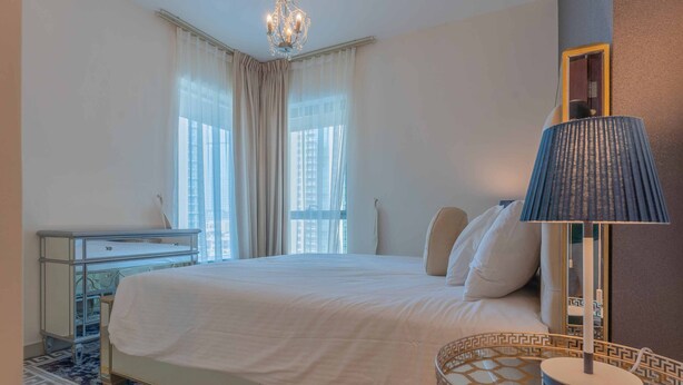 A hotel room with a bed, a bedside table with a lamp, a dresser, and a view of buildings through the window. - Bella Vista - 29 Boulevard Downtown - 2BR - Partial Fountain View (Dubai)