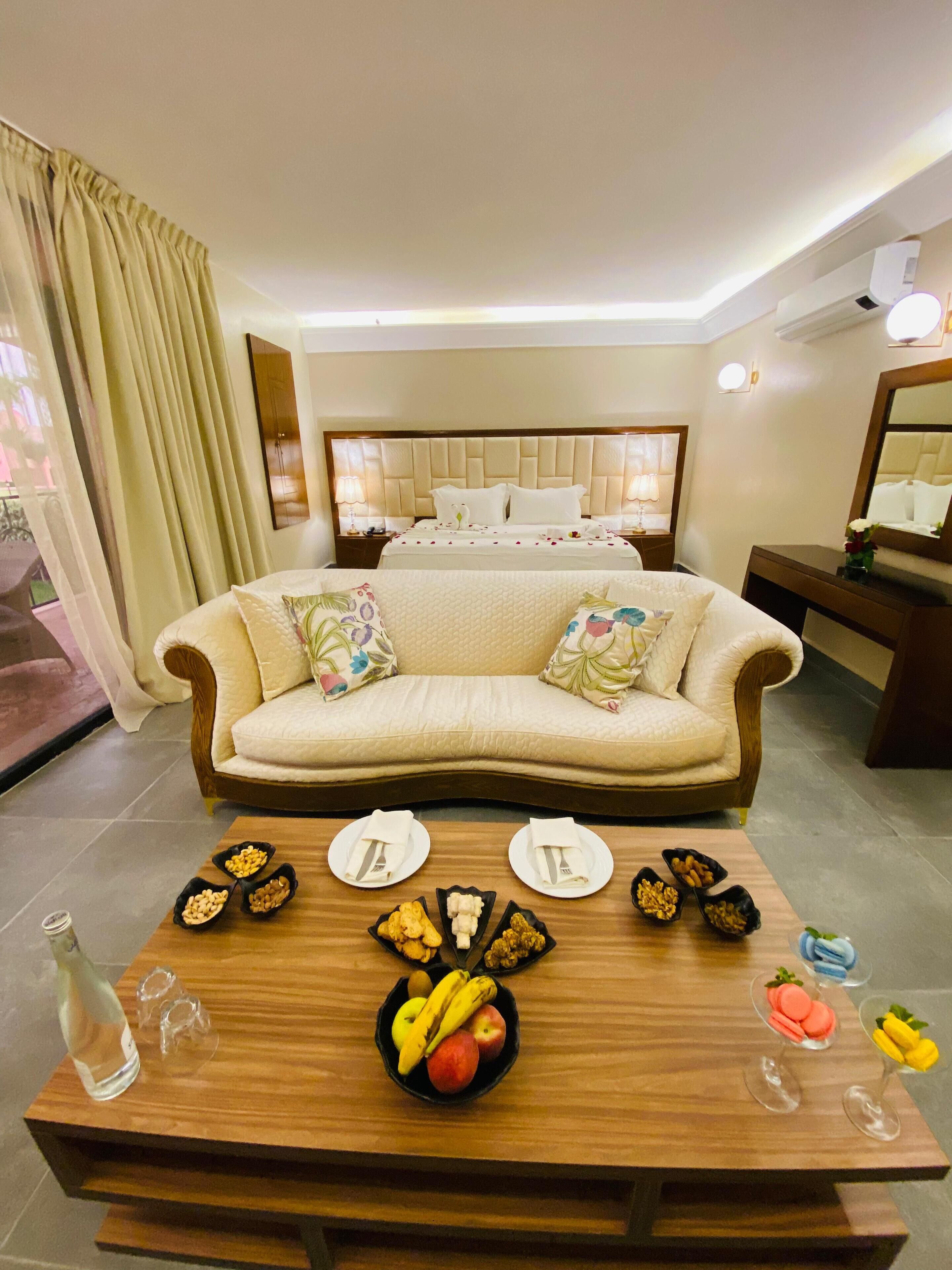 A hotel room with a sofa, a dining table with plates and fruits, a bed with a headboard, and a mirror.
