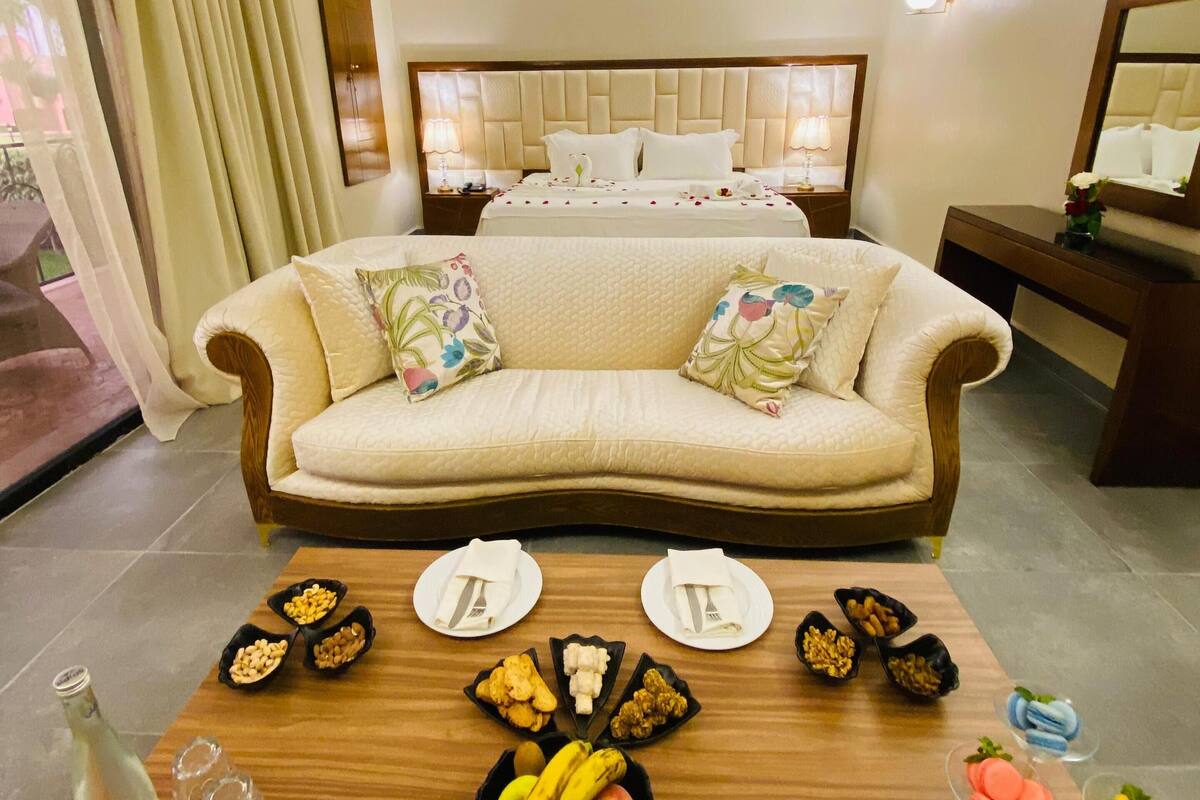 Ein Hotelzimmer mit einer Couch, einem Esstisch mit Tellern und Früchten, einem Bett mit Kopfteil und einem Spiegel.