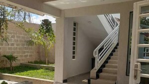 Un espacio exterior techado con piso de concreto, una escalera con barandilla blanca y una puerta con paneles de vidrio.