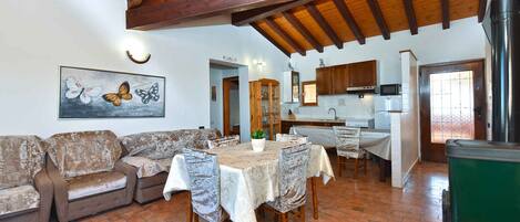 A rustic interior with a wooden ceiling, a dining area with a table and chairs, a kitchen with cabinets and appliances, and a living area with a sofa and a butterfly painting.