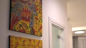 A room with two colorful paintings on the wall, a door, and a glimpse of a living area with a couch and a plant. - Synathens II - Apartments & Studios (Athens)