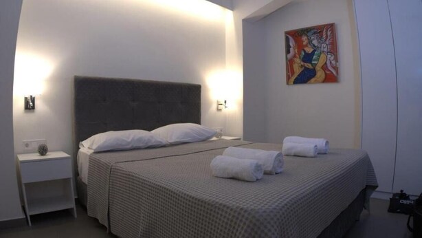 A neatly made bed with a grey headboard and white pillows, a small bedside table with a lamp, and a painting on the wall. - Synathens II - Apartments & Studios (Athens)