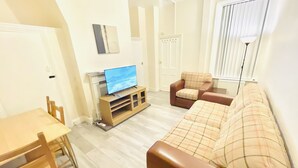 Ein Wohnzimmer mit Fernseher auf einem Holzgestell, einem braunen Sofa, einem karierten Sessel und einem Esstisch mit Stühlen.