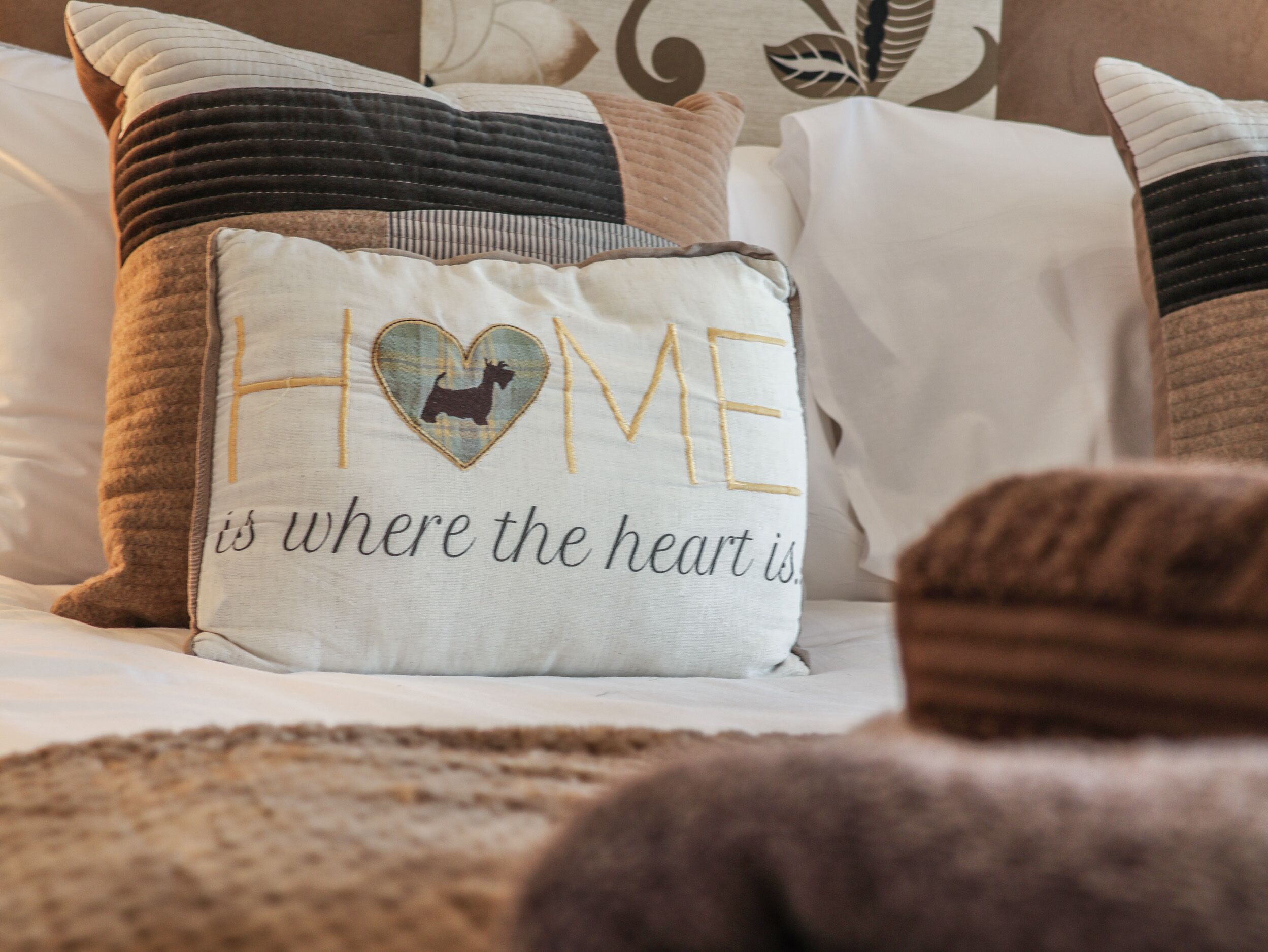 Una cama con varias almohadas, una de ellas con un diseño de corazón y la inscripción 'HOME is where the heart is'.