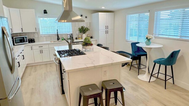 A modern kitchen with white cabinets, a central island, bar stools, and a dining area with a table and chairs. - No Tax! Modern 4BR Pool Villa, Hot Tub, Arcade (Anaheim)