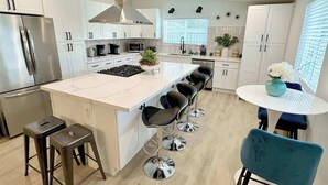 A modern kitchen with white cabinets, a stainless steel refrigerator, a central island with bar stools, and a round table with a vase. - No Tax! Modern 4BR Pool Villa, Hot Tub, Arcade (Anaheim)