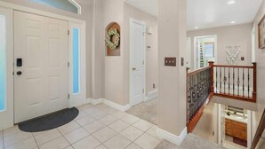 A hallway with a white door, a doormat, a wreath on the wall, and a staircase with a railing.