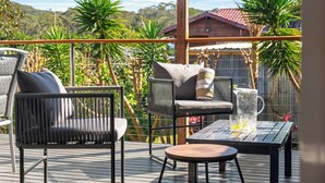 Eine Holzterrasse mit Gartenmöbeln, darunter ein Tisch und Stühle, und Blick auf ein Haus sowie Grünflächen im Hintergrund.