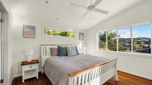 Ein Schlafzimmer mit Bett, Nachttischen, Deckenventilator und großen Fenstern mit Blick auf eine Wohngegend.