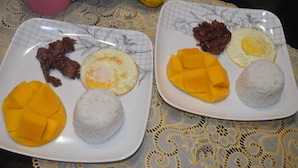 Zwei Teller mit Essen, darunter Mango, Reis und Eier, auf einem Tisch mit weißer Tischdecke.