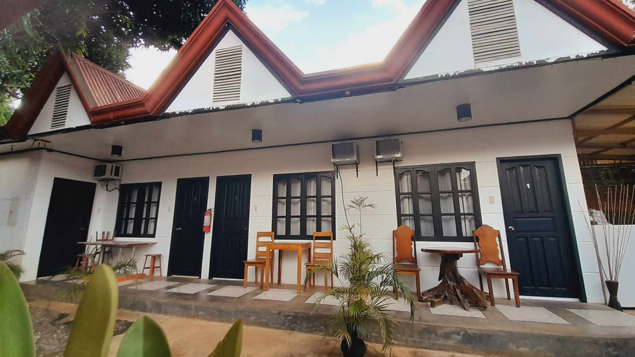 A building with a sloped roof, multiple black doors, and windows with white frames. There are outdoor tables and chairs, and potted plants.