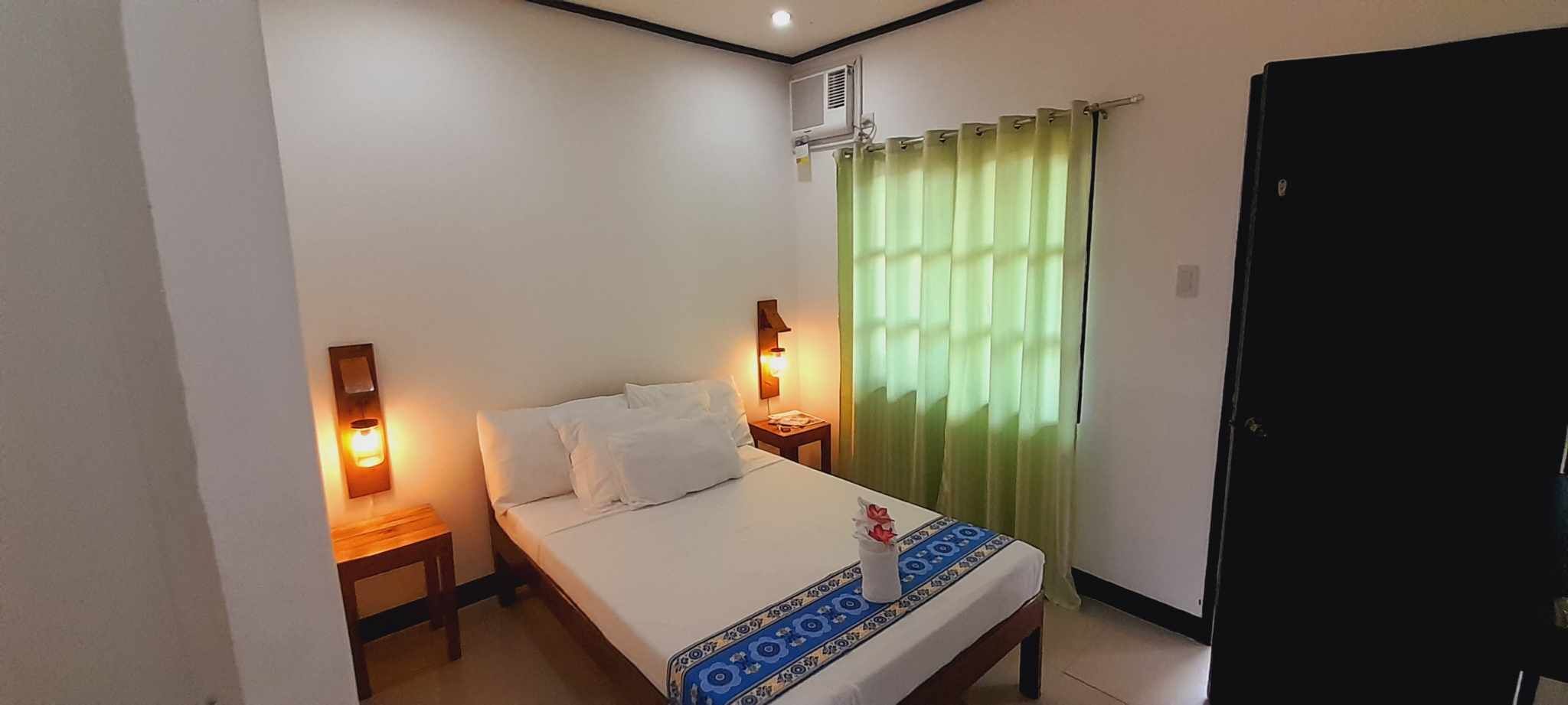 Habitación de hotel con cama, dos mesitas de noche con lámparas, una ventana con cortinas verdes y una puerta cerrada.