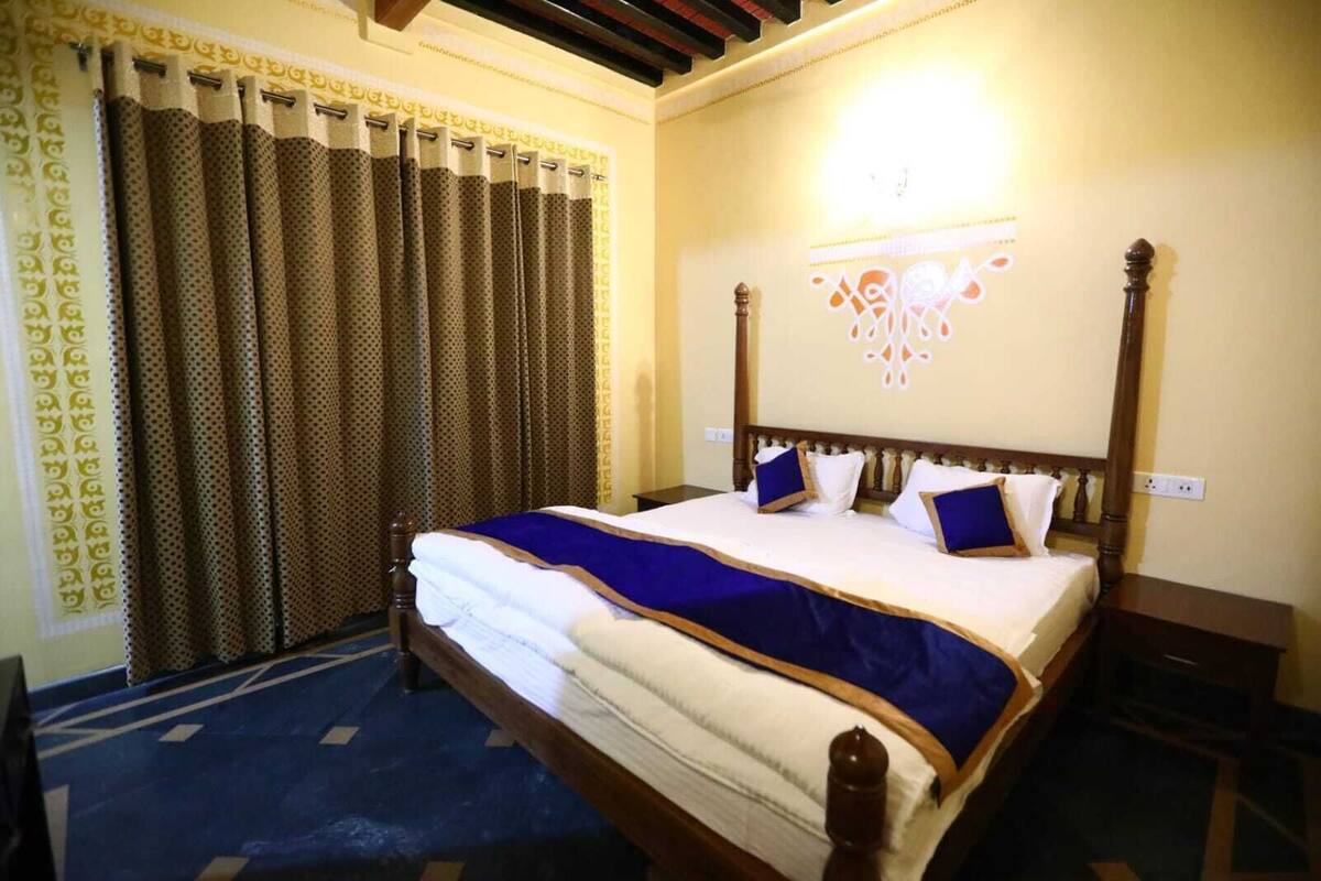 Una cama bien hecha con una colcha azul y blanca, cabecera de madera y cortina estampada.