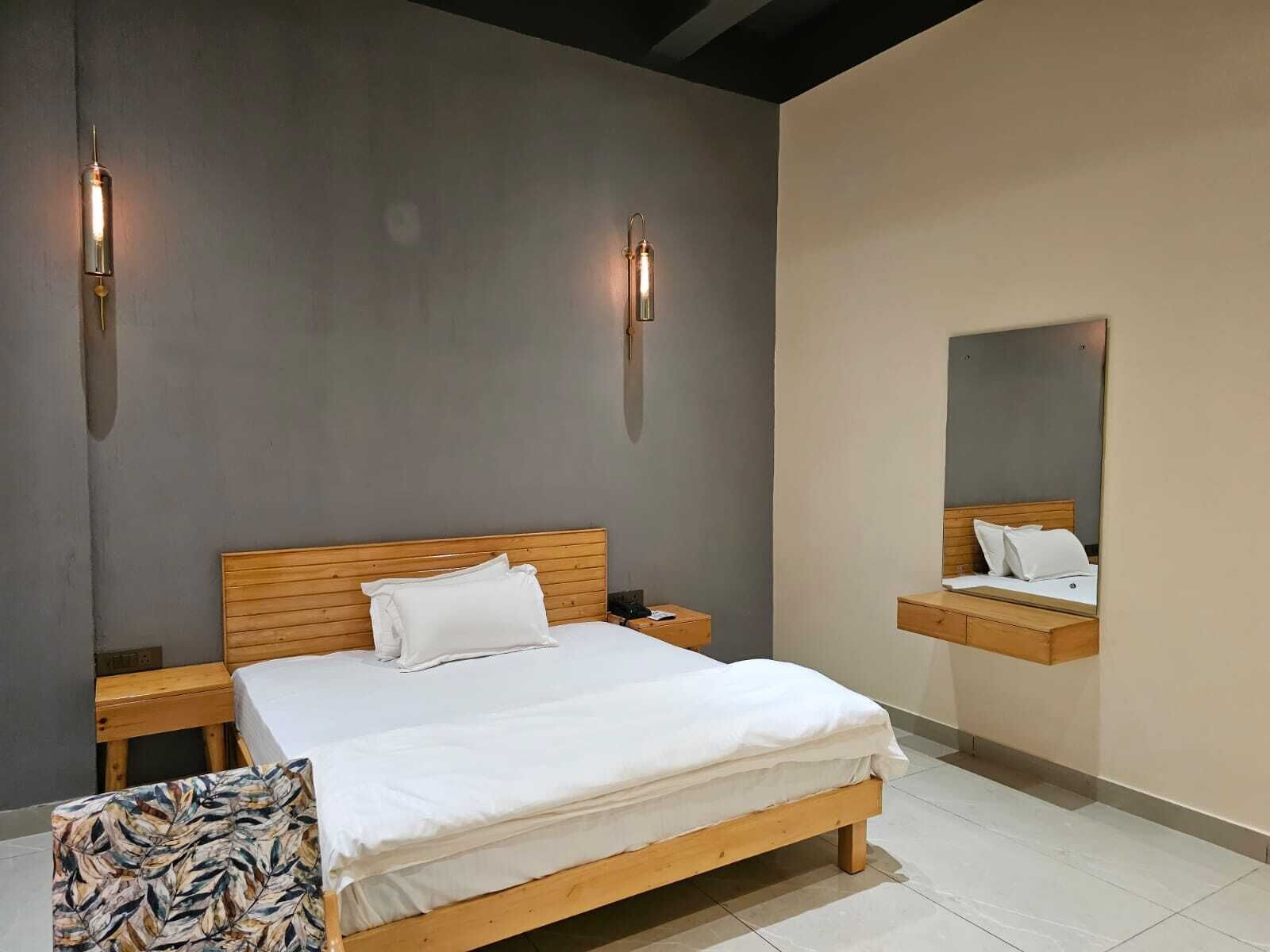 A bedroom with a wooden bed, white bedding, a nightstand, a bedside table, a chair, and two wall-mounted lights.