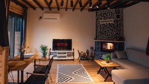 Ein gemütliches Wohnzimmer mit Holzboden, Kamin, einer Couch, einem Fernseher und einer Essecke mit Stühlen und Tisch.