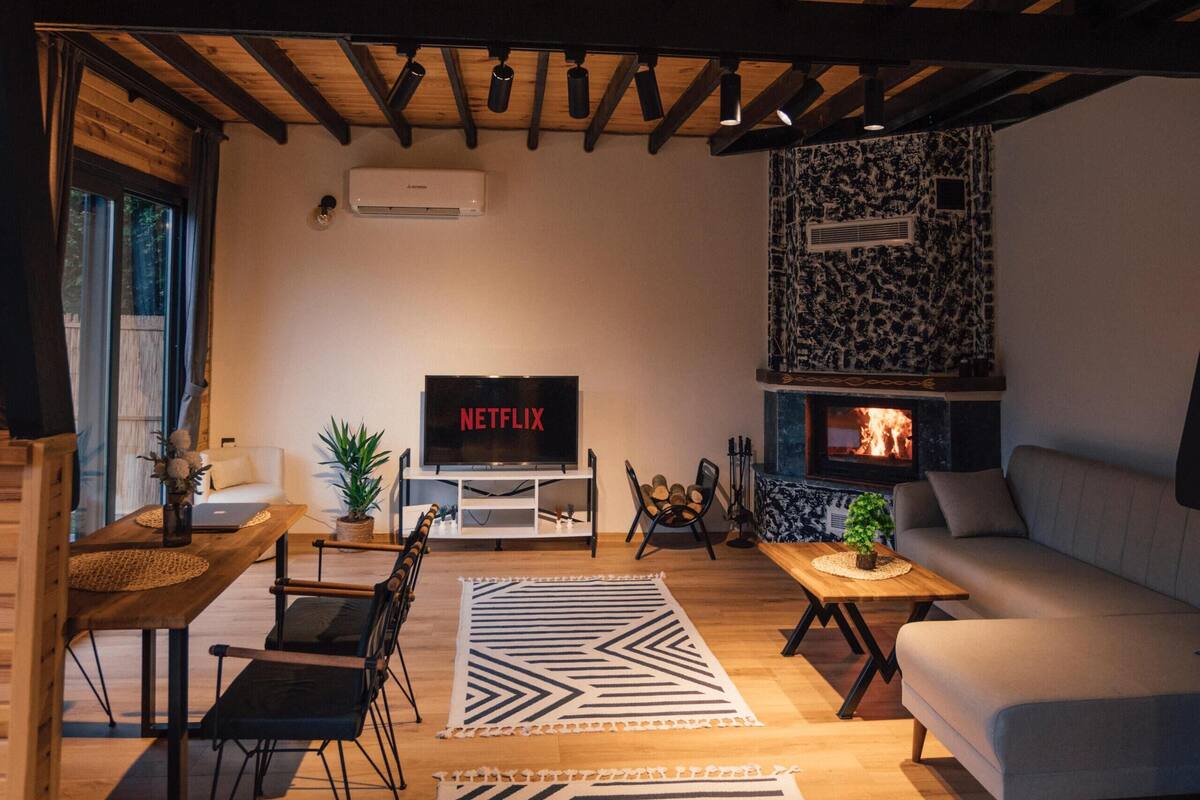 Ein gemütliches Wohnzimmer mit Holzboden, Kamin, einer Couch, einem Fernseher und einer Essecke mit Stühlen und Tisch.