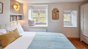 A bedroom with a bed, two windows, a bedside table, a wall-mounted mirror, and a bookshelf. - Stunning 3 bed Yorkshire Dales cottage (Skyreholme)