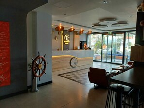 A hotel reception area with a wooden reception desk, a ship's wheel decoration, and a large window offering a view of the street outside. - Del Puerto Hotel (Aliaga)