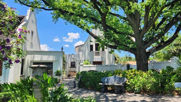 A white building with a large tree in front, a stone bench, and a 'WELCOME' sign. - Oak View Manor (Lesedi)
