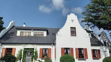 A white building with a steeply pitched roof, red shutters, and a small garden with a neatly trimmed tree.