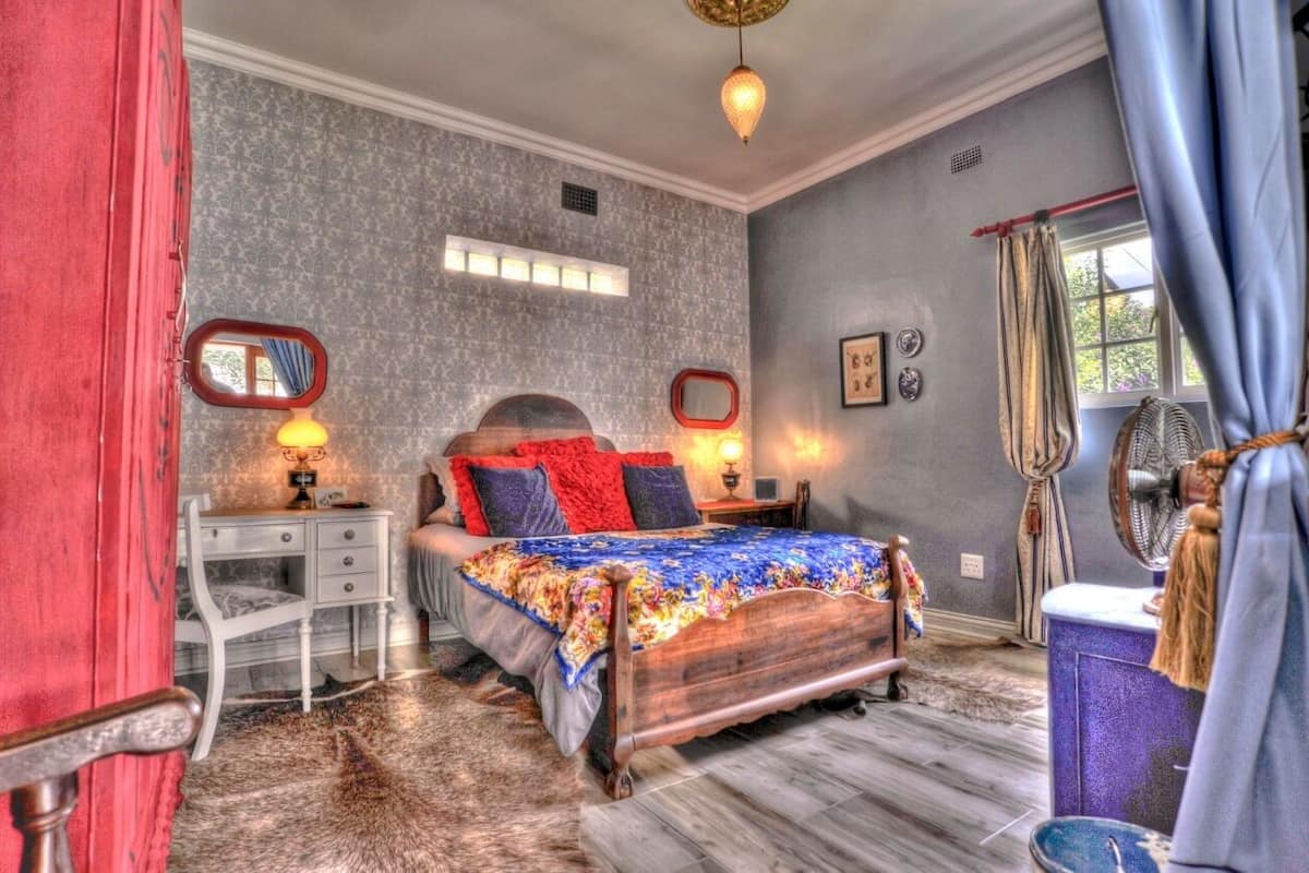 Quarto com cama de madeira, duas mesas de cabeceira com abajures, cômoda, espelho e janela com cortinas.