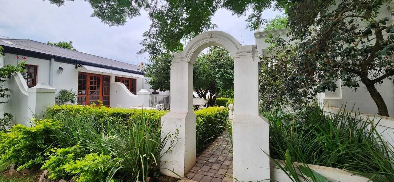 A white building with a red door, surrounded by lush greenery and a stone pathway leading through an arched entrance.