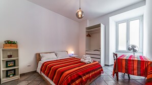 A bedroom with a bed, a bedside table with a lamp, a wooden shelf with pictures, a window with a fan, and a table with a striped tablecloth. - Casa Fabrizio (Rome)