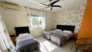 Une chambre avec deux lits, un ventilateur de plafond, une fenêtre avec des rideaux, une chaise et un mur orné d’un motif de feuilles.