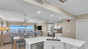 A modern kitchen with a dishwasher, white cabinets, and a large island with a sink. There is a dining area with a table and chairs, and a view of the ocean through large windows. - Admirals Quarters 1403-Beachfront | 2 King Suites | Free parking for 2 Vehicles (Orange Beach)