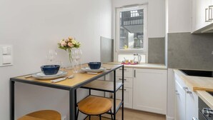 Une cuisine compacte avec des meubles blancs, une petite table à manger agrémentée de bols bleus et de verres à vin, et une fenêtre donnant sur la ville.