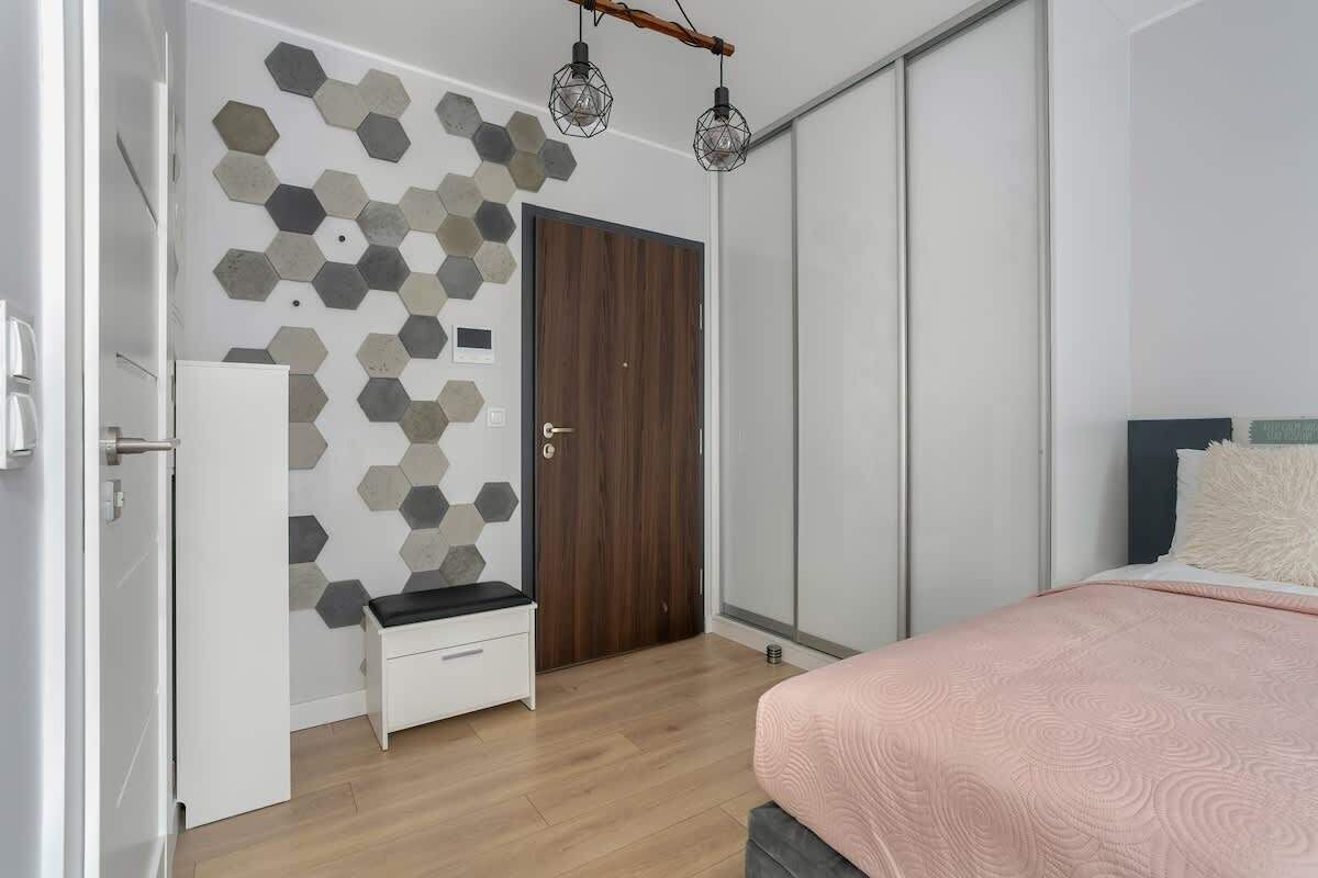 A bedroom with a wooden wardrobe, a white bedside table, a bed with a pink blanket, and a hexagonal tile wall.