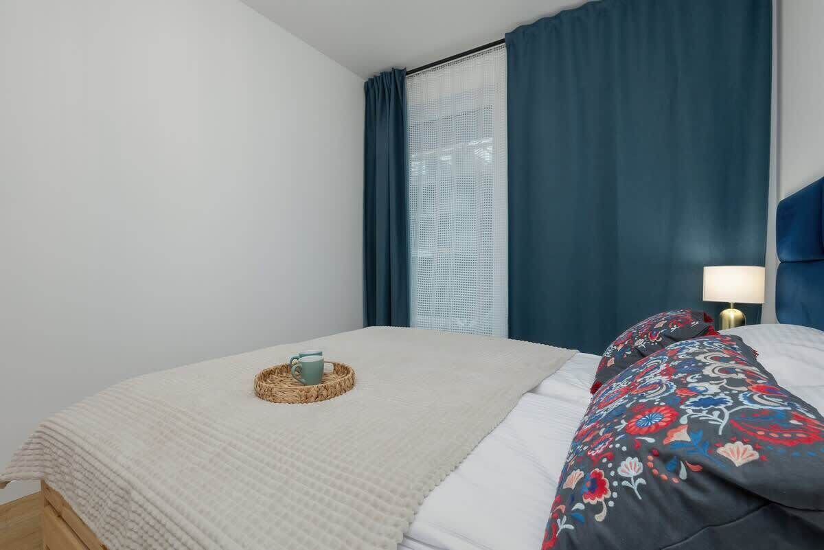 A neatly made bed with a white comforter and a decorative pillow, a woven basket with a cup, and a bedside lamp on a wooden nightstand.