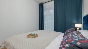 A neatly made bed with a white comforter and a decorative pillow, a woven basket with a cup, and a bedside lamp on a wooden nightstand.