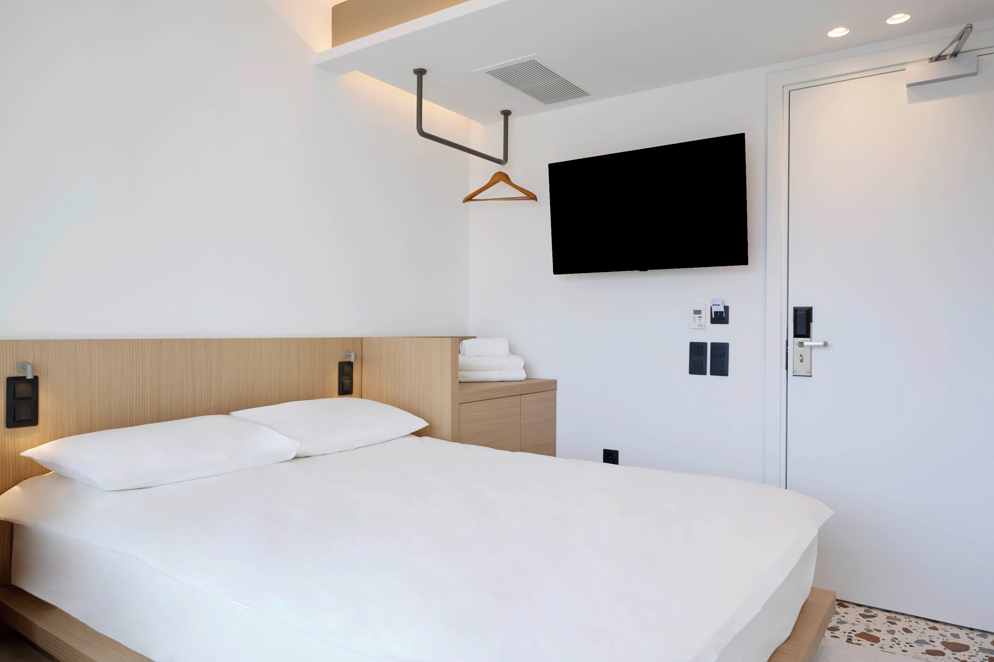 A modern hotel room with a large bed, a mounted TV, a wooden headboard, a bedside table with a towel, and a door with a handle.