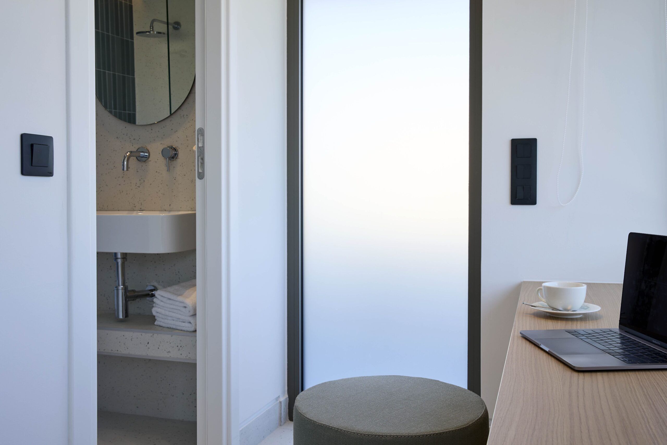 Un baño moderno con lavamanos, espejo y toallero. Hay un ordenador portátil sobre una mesa de madera con una taza y un plato.
