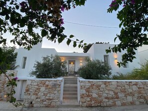 A white building with a stone entrance, steps leading up to a door, and a small garden area with shrubs and a tree. - Nisides Villas Naoussa (Naousa)