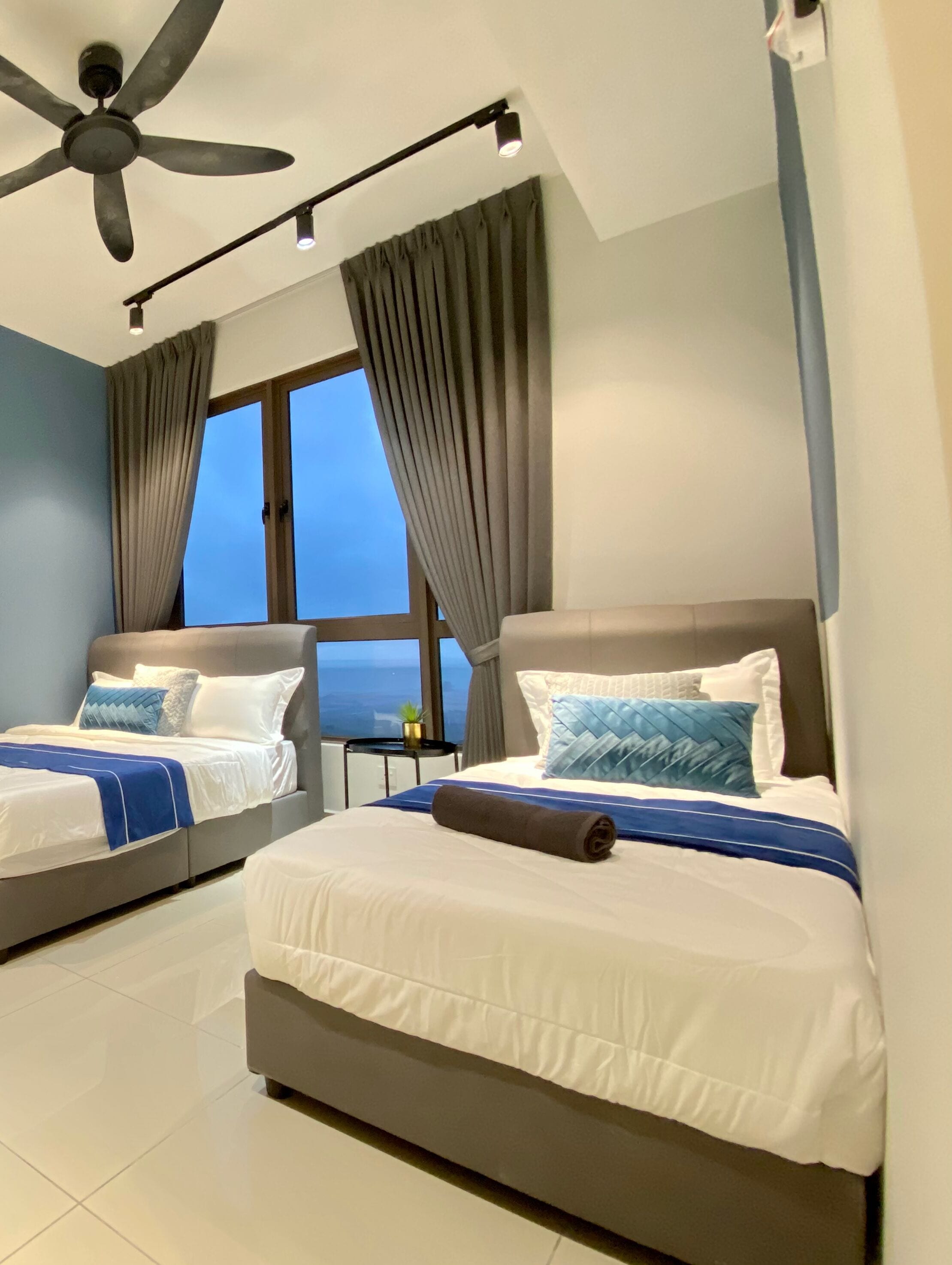 A hotel room with two beds, a ceiling fan, a small table with a plant, and a view of the ocean through a large window.