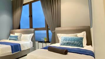 A hotel room with two beds, a ceiling fan, a small table with a plant, and a view of the ocean through a large window.