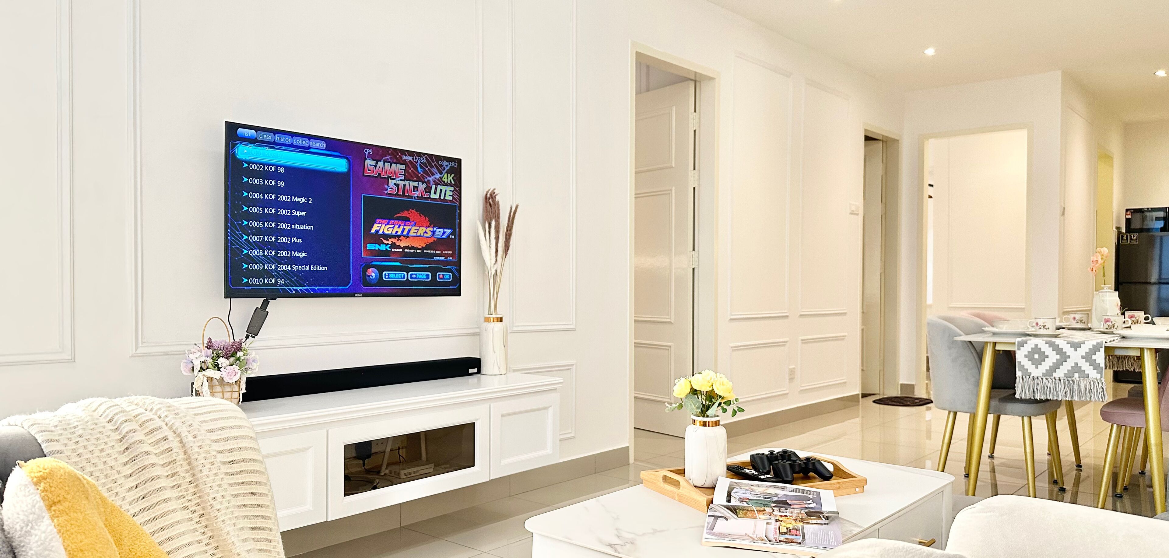 A modern living room with a flat-screen TV mounted on the wall, a white entertainment unit, a coffee table with magazines, and a dining area with a table and chairs.