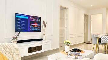 A modern living room with a flat-screen TV mounted on the wall, a white entertainment unit, a coffee table with magazines, and a dining area with a table and chairs.