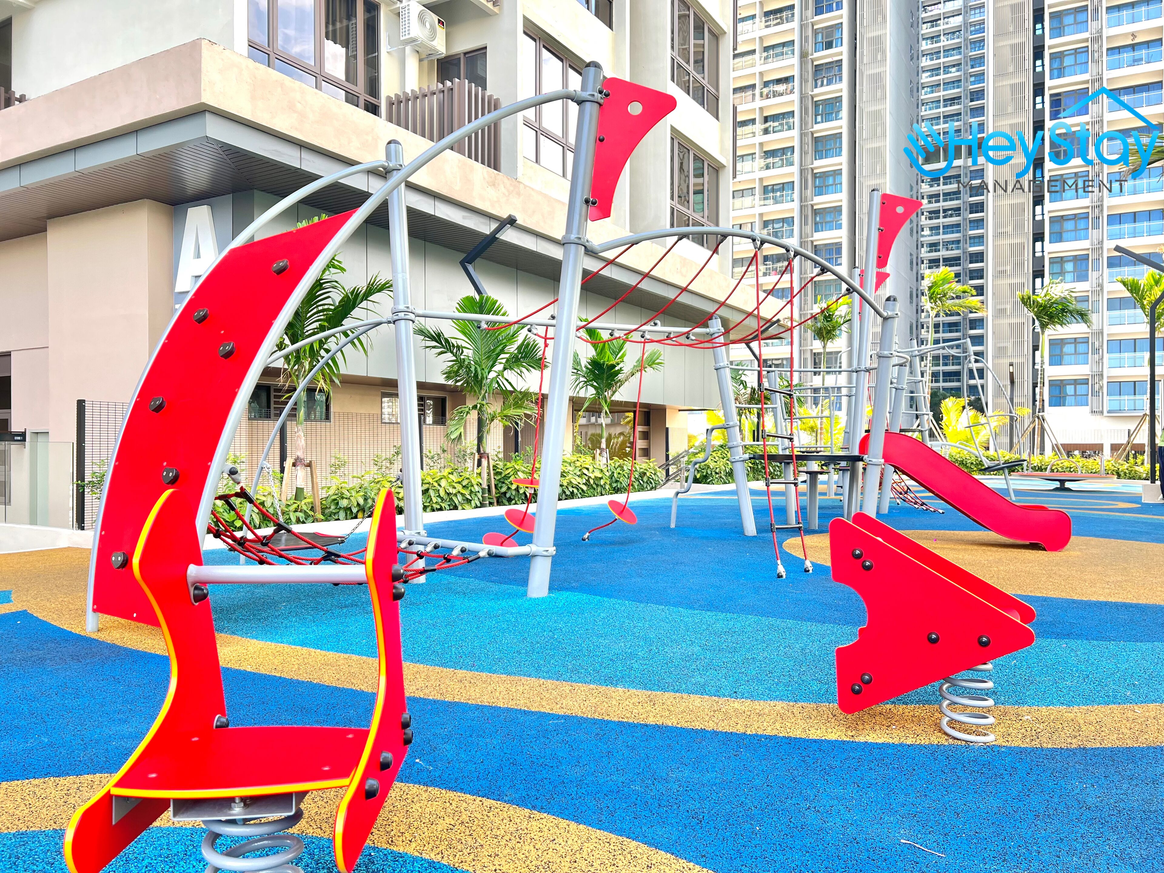 A playground with red and yellow equipment, surrounded by a blue and yellow play area, and buildings in the background.