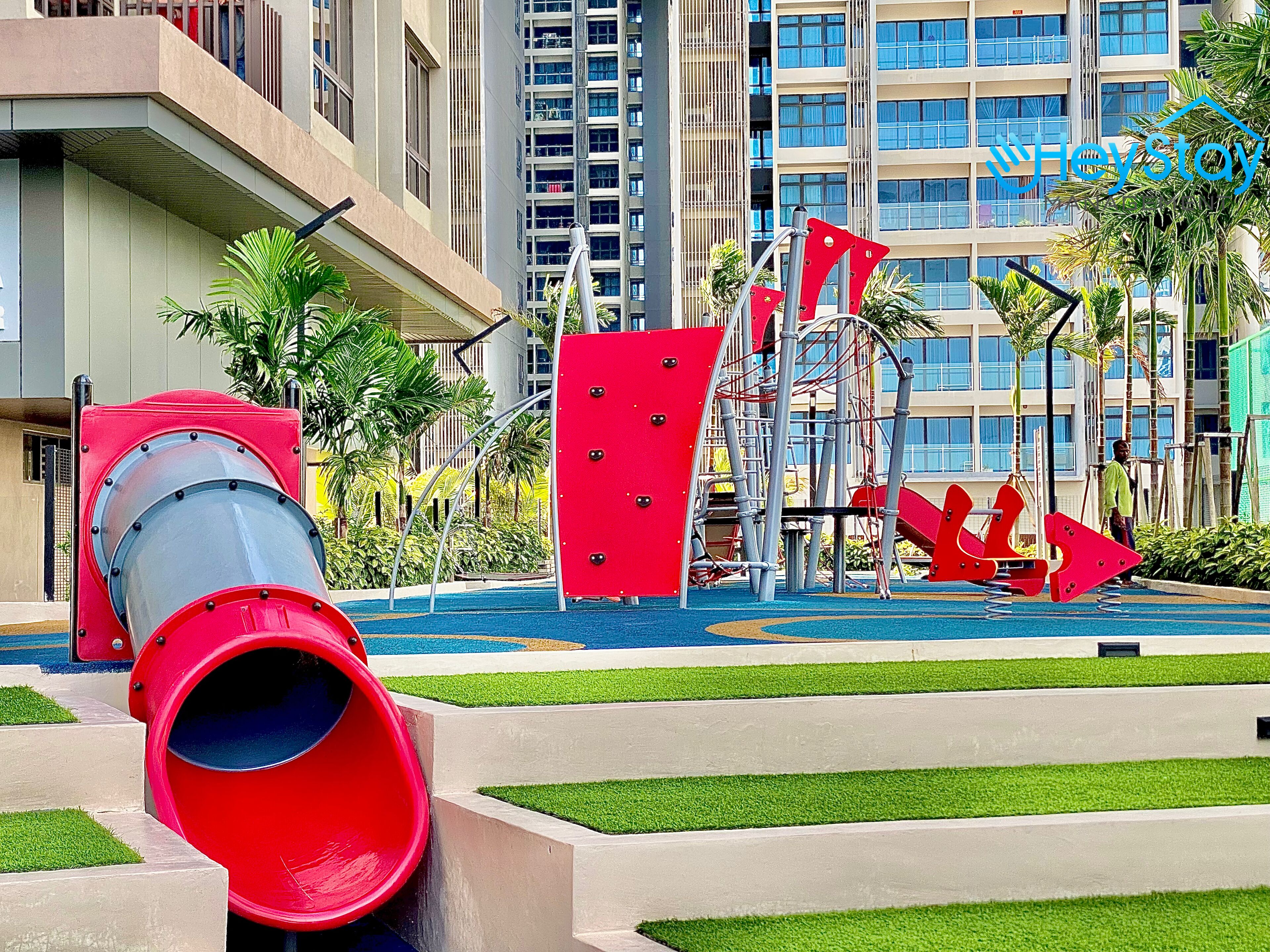 A playground with a red slide and climbing structures, surrounded by palm trees and a building with multiple balconies.