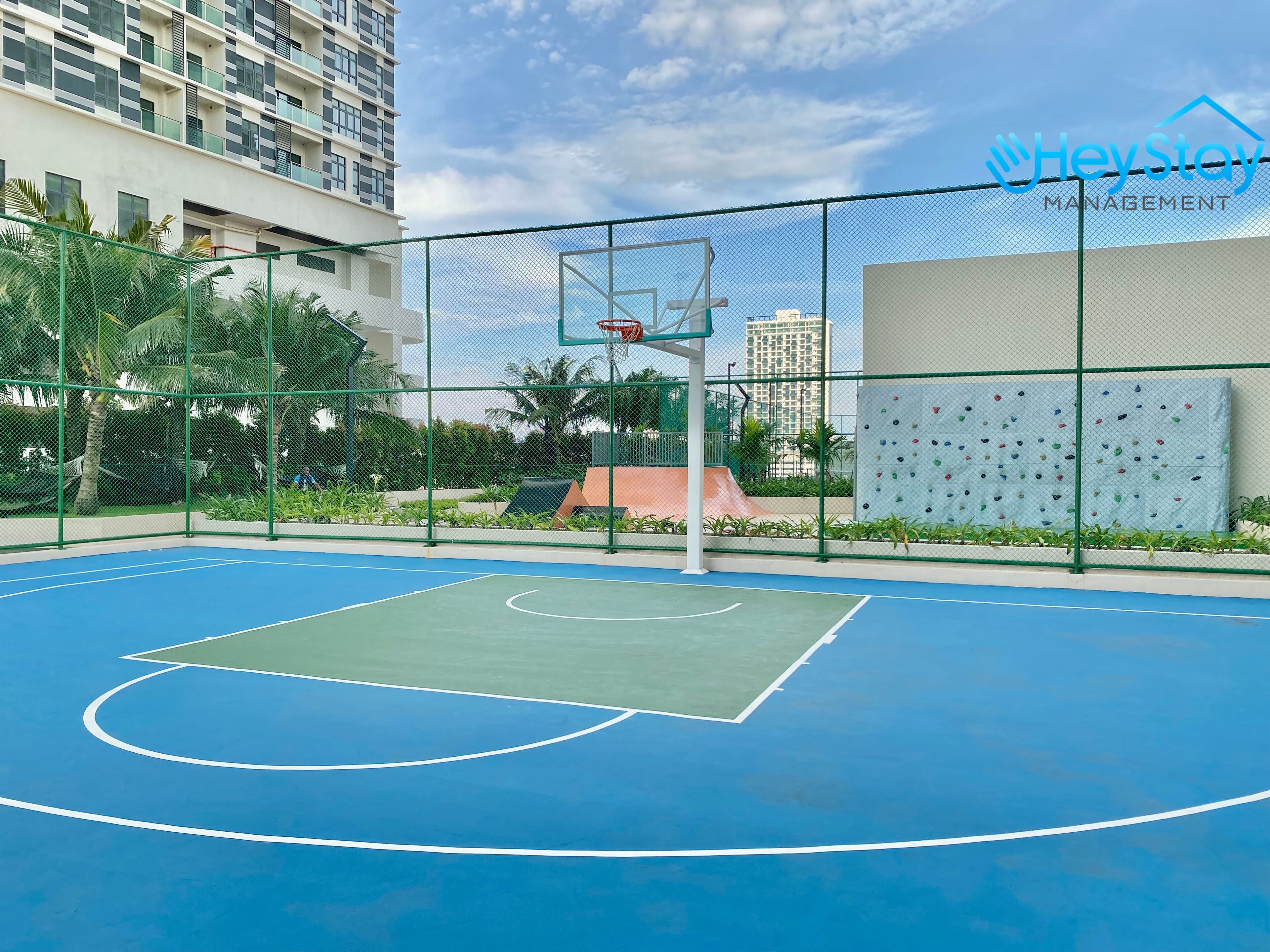 Un terrain de basket en plein air, entouré d’une clôture verte, avec un panier de basket et un mur d’escalade.
