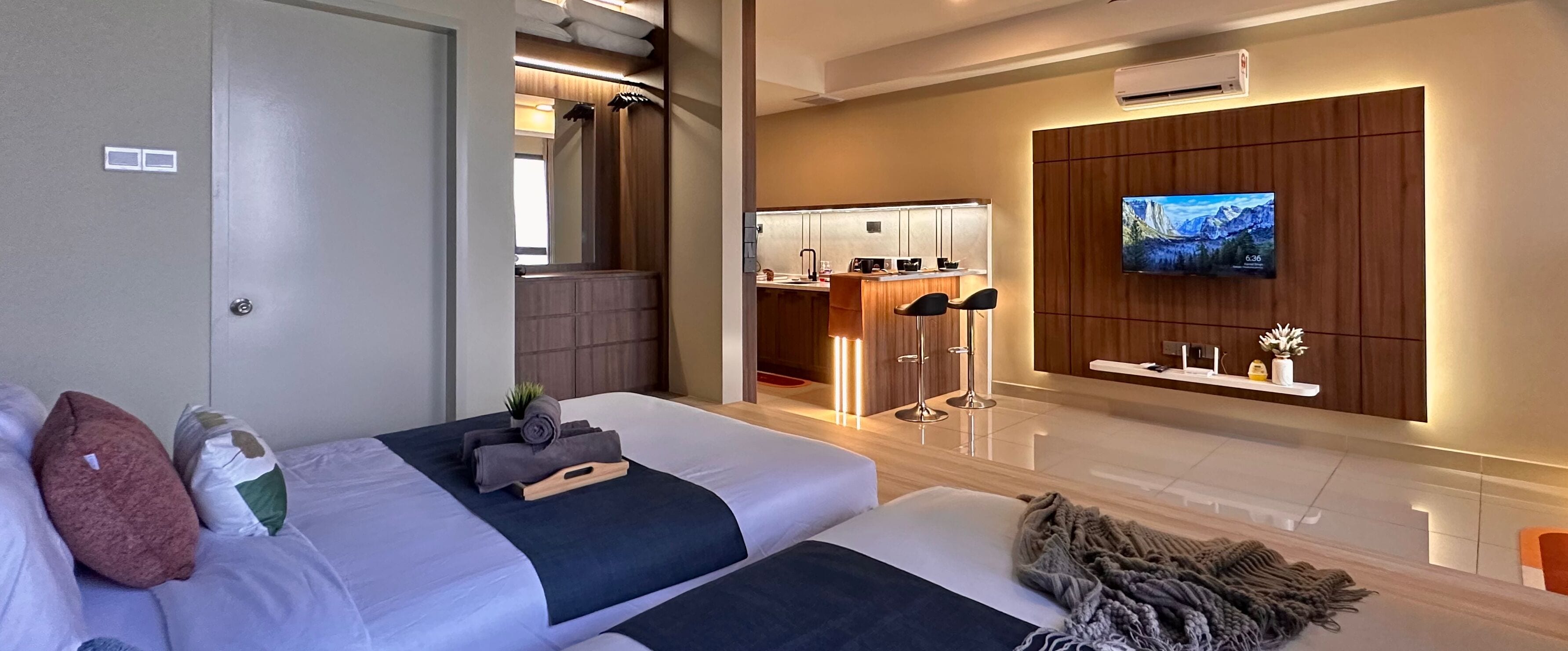 A modern hotel room with a large bed, a TV mounted on the wall, a small kitchen area, and a bathroom visible through an open door.