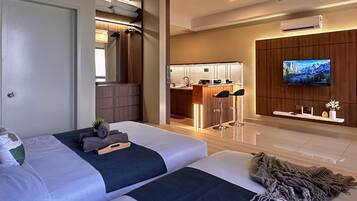 A modern hotel room with a large bed, a TV mounted on the wall, a small kitchen area, and a bathroom visible through an open door.