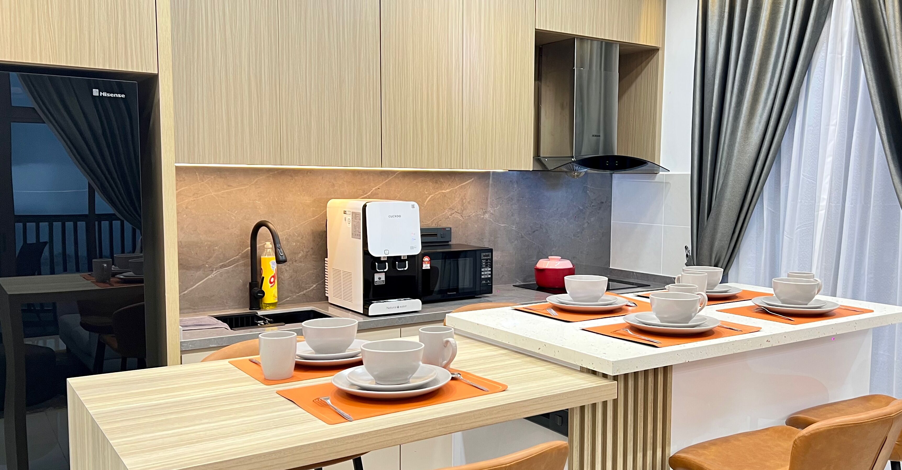 Una cocina moderna con cafetera integrada, microondas y estufa. Hay dos mesas de comedor con individuales anaranjados y tazas blancas.